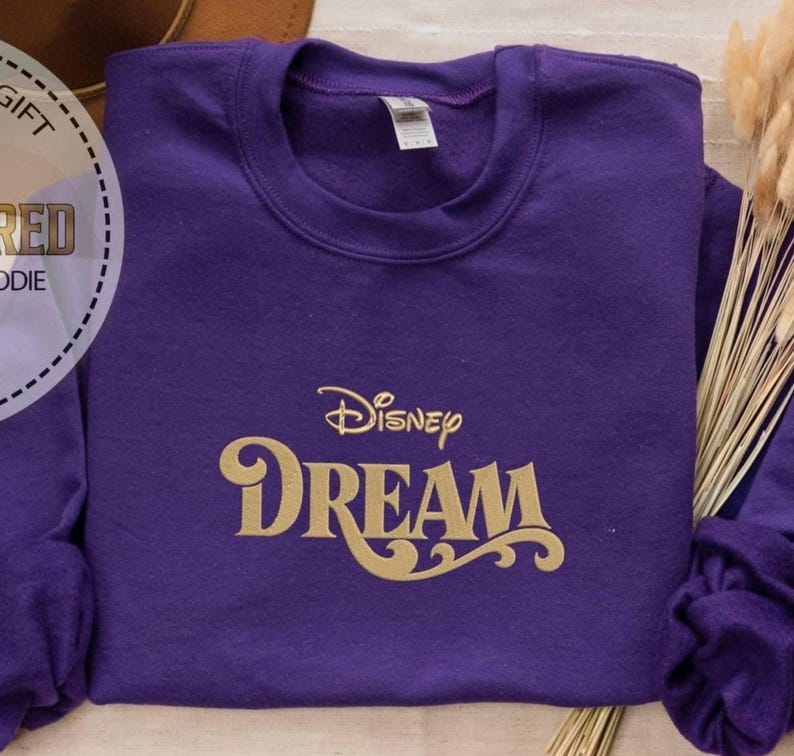 Peut inclure: Un sweat-shirt violet avec l'inscription dor&eacute;e "Disney DREAM". Le sweat-shirt &agrave; col rond est pli&eacute;. L'image est d'un v&ecirc;tement.