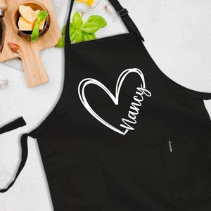 May include: Black apron with a white heart and the name "Nancy" written inside. The apron has a pocket on the front.