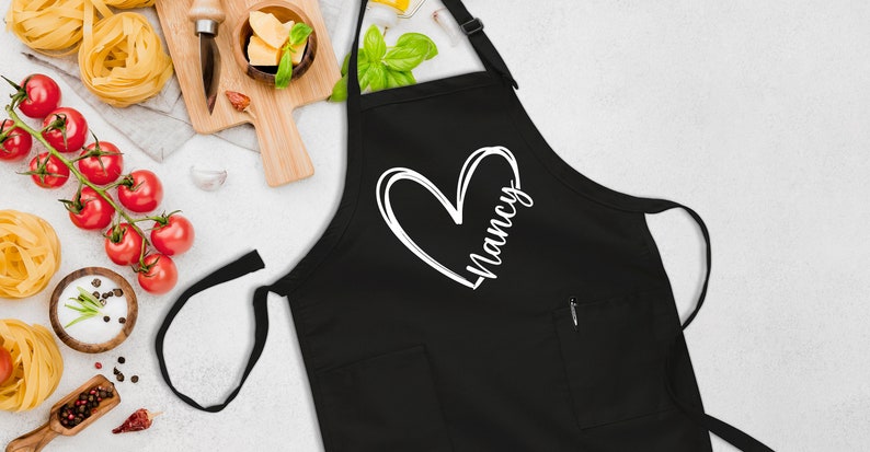 May include: Black apron with a white heart and the name "Nancy" written inside. The apron has a pocket on the front.