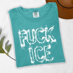 May include: Teal Comfort Colors t-shirt with the words "FUCK ICE" in distressed white lettering. A brown hat is partially visible in the background. The shirt is folded and lies on a white surface.