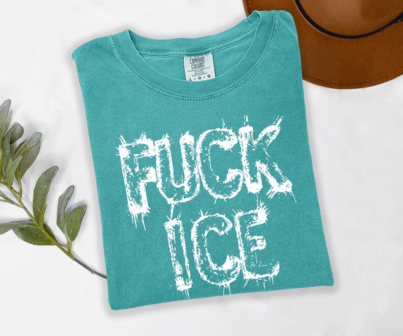 May include: Teal Comfort Colors t-shirt with the words "FUCK ICE" in distressed white lettering. A brown hat is partially visible in the background. The shirt is folded and lies on a white surface.