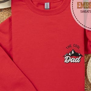 Op de afbeelding: Een rode sweatshirt met de geborduurde tekst "THE COOL Dad" boven een bergafbeelding. De sweatshirt is gemaakt van zacht materiaal en is netjes opgevouwen. Het Gildan-merklabel is zichtbaar op de kraag.