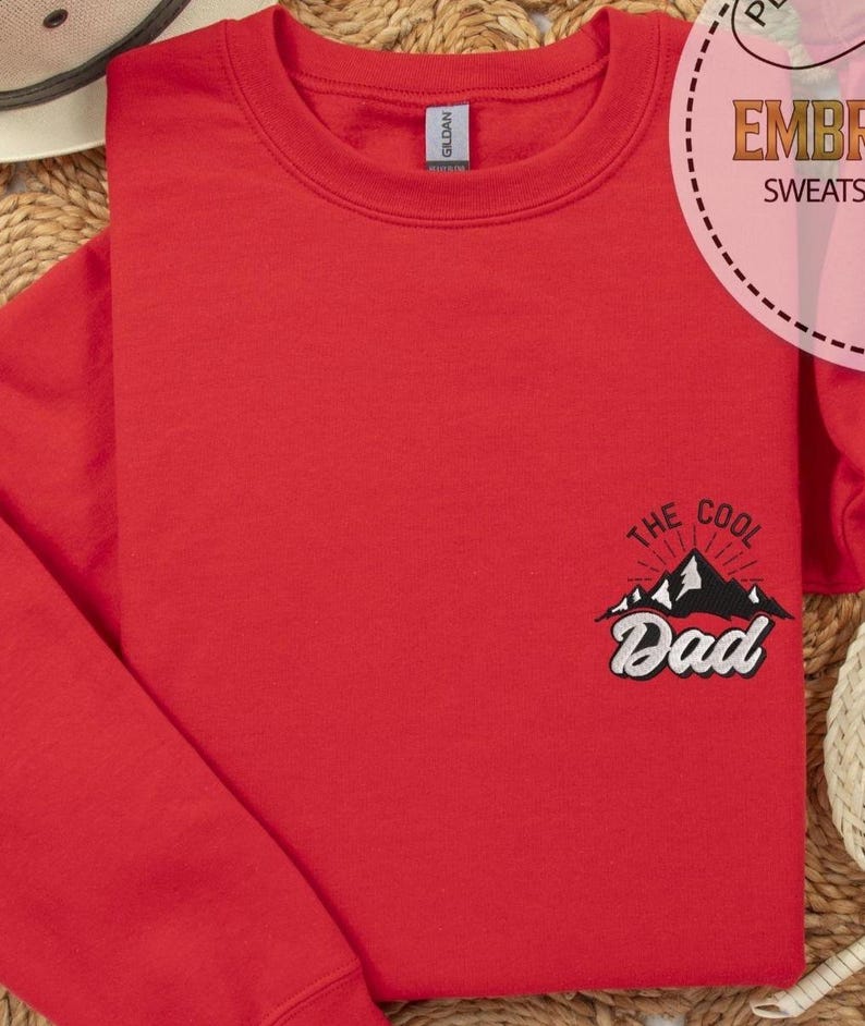 Op de afbeelding: Een rode sweatshirt met de geborduurde tekst "THE COOL Dad" boven een bergafbeelding. De sweatshirt is gemaakt van zacht materiaal en is netjes opgevouwen. Het Gildan-merklabel is zichtbaar op de kraag.
