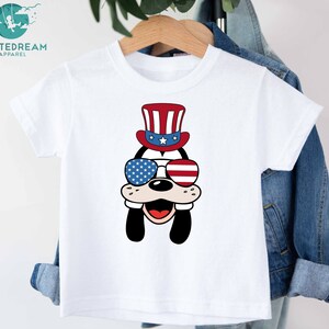 May include: White t-shirt with a cartoon image of a dog wearing a red, white, and blue hat and sunglasses. The dog is smiling and has a red nose. The hat has stars and stripes.
