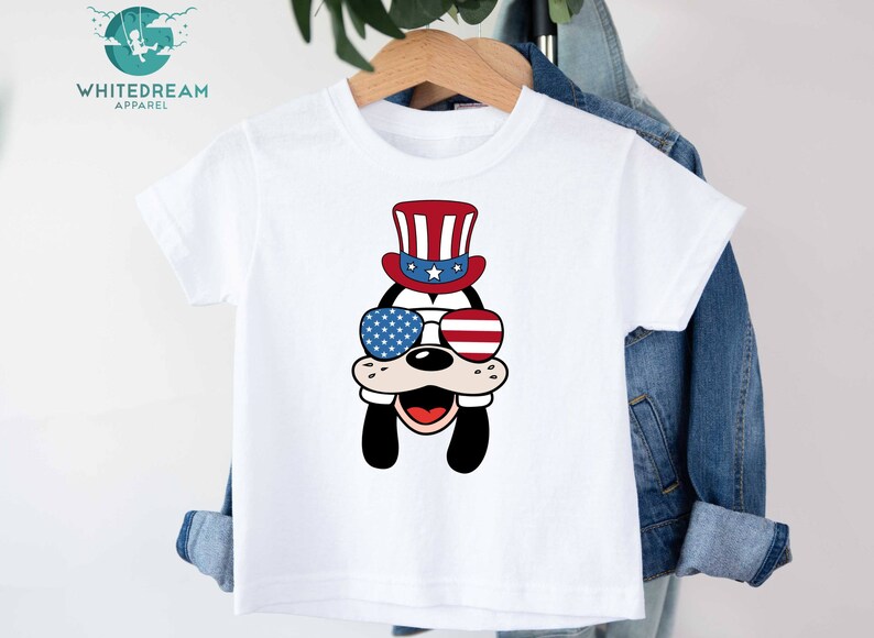May include: White t-shirt with a cartoon image of a dog wearing a red, white, and blue hat and sunglasses. The dog is smiling and has a red nose. The hat has stars and stripes.