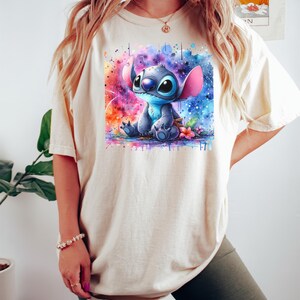 Pu&ograve; includere: T-shirt color crema con un'illustrazione ad acquerello del personaggio Stitch di Lilo & Stitch. L'opera mostra Stitch in blu con grandi orecchie, su uno sfondo vibrante e colorato con spruzzi di rosa, blu e arancione.