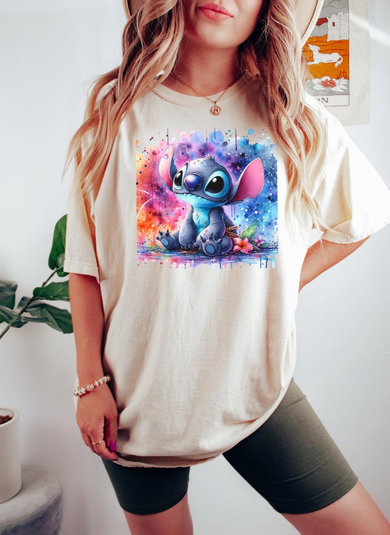 Pu&ograve; includere: T-shirt color crema con un'illustrazione ad acquerello del personaggio Stitch di Lilo & Stitch. L'opera mostra Stitch in blu con grandi orecchie, su uno sfondo vibrante e colorato con spruzzi di rosa, blu e arancione.