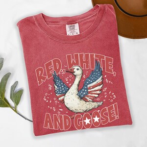 May include: Red t-shirt with a graphic of a goose with wings designed as the American flag. The text "RED WHITE AND GOOSE!" is printed below the goose. The shirt is a Comfort Colors brand.