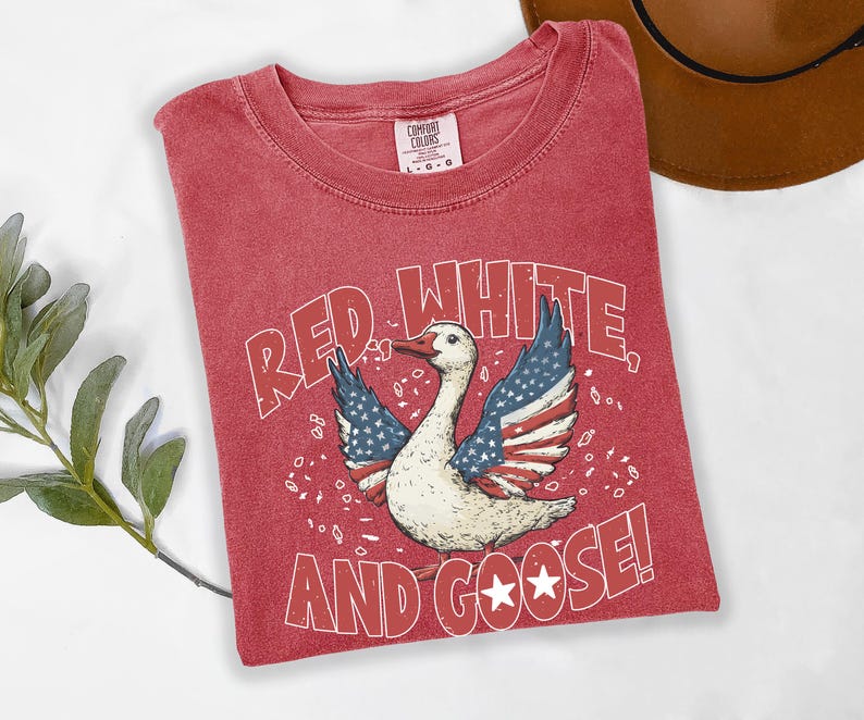 May include: Red t-shirt with a graphic of a goose with wings designed as the American flag. The text "RED WHITE AND GOOSE!" is printed below the goose. The shirt is a Comfort Colors brand.