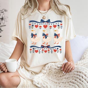 May include: Cream-colored t-shirt featuring a whimsical design of three geese adorned with patriotic bows. The design includes red hearts, blue bows, and floral accents, creating a charming and festive aesthetic. The shirt is a relaxed fit.