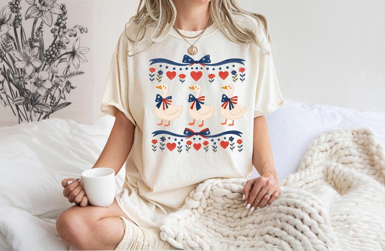 May include: Cream-colored t-shirt featuring a whimsical design of three geese adorned with patriotic bows. The design includes red hearts, blue bows, and floral accents, creating a charming and festive aesthetic. The shirt is a relaxed fit.