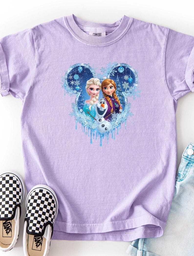 Puede incluir: Una camiseta de color lila claro con un dise&ntilde;o de Elsa, Anna y Olaf de la pel&iacute;cula Frozen, enmarcado en un dise&ntilde;o con forma de Mickey Mouse con copos de nieve. La camiseta se muestra con zapatos sin cordones a cuadros blancos y negros y pantalones cortos vaqueros.