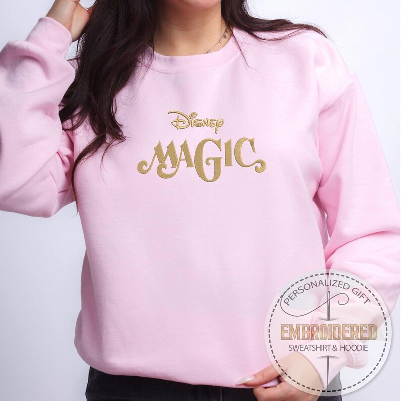 Peut inclure: Sweat-shirt rose clair avec le mot "Disney" brod&eacute; en or au-dessus de "MAGIC". Le sweat-shirt a des manches longues et un col rond. Une &eacute;tiquette circulaire avec le texte "PERSONALIZED GIFT EMBROIDERED SWEATSHIRT & HOODIE" est en bas &agrave; droite.