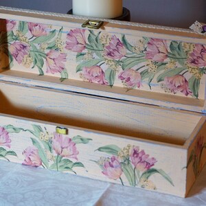 May include: A rectangular wooden box with a floral design on the lid and sides. The box is painted in a light beige color with a distressed finish. The floral design features pink tulips and green leaves.
