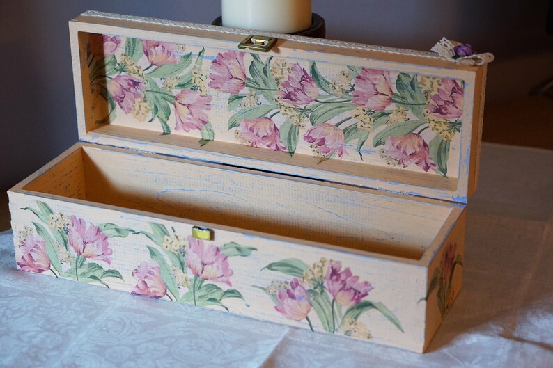May include: A rectangular wooden box with a floral design on the lid and sides. The box is painted in a light beige color with a distressed finish. The floral design features pink tulips and green leaves.