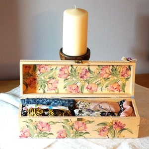 May include: A wooden box with a floral design and a light pink and yellow color scheme. The box is open and contains several small fabric items.