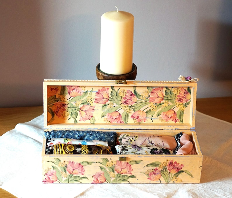 May include: A wooden box with a floral design and a light pink and yellow color scheme. The box is open and contains several small fabric items.