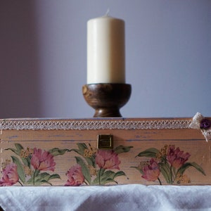 May include: A pink wooden box with a floral design and a white lace trim. The box has a brass latch and a small flower embellishment on the top right corner. The box is sitting on a white tablecloth in front of a white candle on a wooden stand.