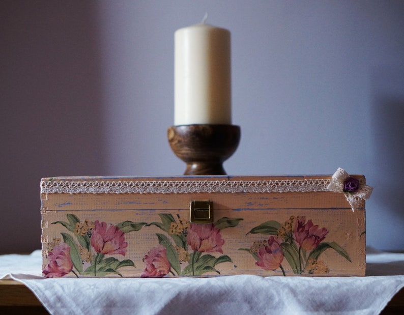May include: A pink wooden box with a floral design and a white lace trim. The box has a brass latch and a small flower embellishment on the top right corner. The box is sitting on a white tablecloth in front of a white candle on a wooden stand.