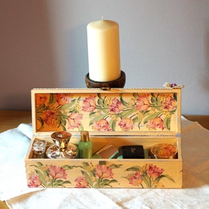 May include: A floral patterned wooden box with a hinged lid is open, revealing a variety of items inside, including a silver bottle stopper, a bottle of liquid, a small box, and a jar. The box is decorated with a floral pattern of pink and yellow tulips.