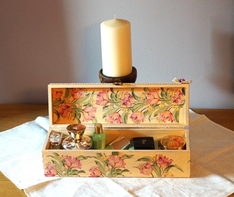 May include: A floral patterned wooden box with a hinged lid is open, revealing a variety of items inside, including a silver bottle stopper, a bottle of liquid, a small box, and a jar. The box is decorated with a floral pattern of pink and yellow tulips.