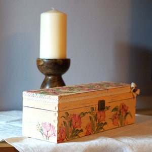 May include: A decorative wooden box with a floral design and a white lace trim. The box is painted in a light pink color and features a floral pattern with pink, green, and white flowers. The box has a hinged lid and a small metal latch.