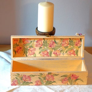 May include: A light yellow wooden box with a floral design on the lid and bottom. The box is open and the inside is empty. The box is sitting on a white cloth.