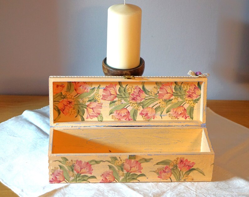 May include: A light yellow wooden box with a floral design on the lid and bottom. The box is open and the inside is empty. The box is sitting on a white cloth.