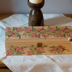 May include: A rectangular wooden box with a floral design featuring pink tulips and green leaves. The box has a white lace trim and a small purple flower embellishment on the side.
