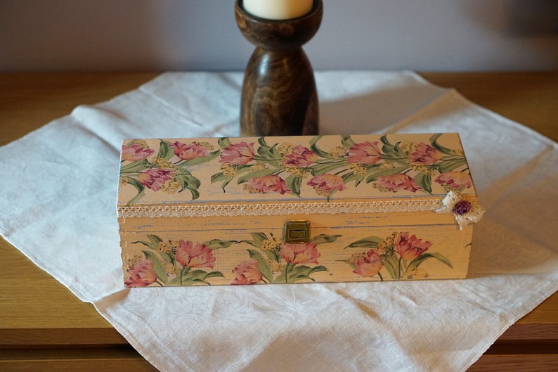 May include: A rectangular wooden box with a floral design featuring pink tulips and green leaves. The box has a white lace trim and a small purple flower embellishment on the side.
