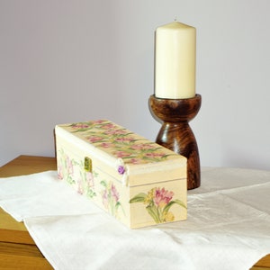 May include: A decorative wooden box with a floral design and a hinged lid. The box is painted in shades of cream, pink, and green. A wooden candle holder with a white candle is in the background.