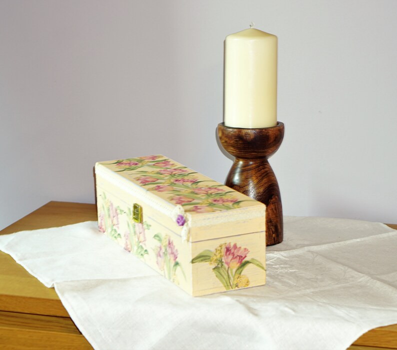 May include: A decorative wooden box with a floral design and a hinged lid. The box is painted in shades of cream, pink, and green. A wooden candle holder with a white candle is in the background.