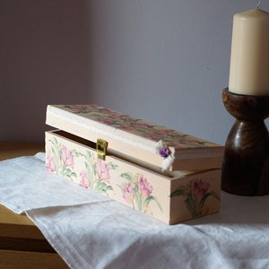 May include: A rectangular wooden box with a floral design painted on the lid and sides. The box is open and has a brass latch. The box is sitting on a white tablecloth on a wooden table.