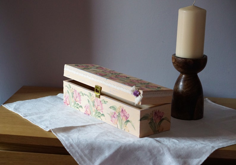 May include: A rectangular wooden box with a floral design painted on the lid and sides. The box is open and has a brass latch. The box is sitting on a white tablecloth on a wooden table.