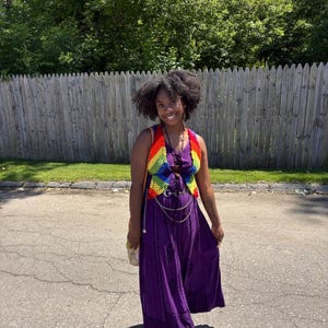Peut inclure: Un gilet sans manches en crochet arborant un motif arc-en-ciel vibrant porté sur une robe longue violette unie. La robe présente un détail de laçage sur le devant, et la tenue est accessoirisée avec une ceinture à chaînes argentée à boucle ronde.
