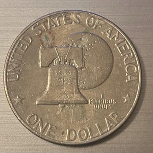 Op de afbeelding: Een zilveren dollar munt met de Liberty Bell en de inscripties "United States of America" en "One Dollar". De munt is cirkelvormig, met sterren en het motto "E Pluribus Unum".