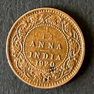 May include: A copper-colored coin with the inscription "1/2 ANNA INDIA 1920" surrounded by a decorative border of floral designs.