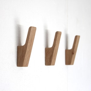 Op de afbeelding: Drie lichtbruine houten wandhaken met een gladde afwerking en een schuin, omhoog gericht ontwerp. Elke haak is afzonderlijk gemonteerd op een effen witte muur, wat de natuurlijke houtnerftextuur en een eenvoudige, functionele vorm laat zien.