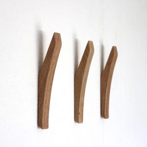 以下が含まれることがあります： 湾曲した細長いデザインのライトブラウンの木製ウォールフック3つ。各フックは滑らかな天然木の木目調で、壁から外側に向かって垂直に取り付けられており、物を掛けるための機能的なポイントを提供しています。