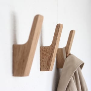 May include: Three light brown wooden wall hooks with a natural grain texture. Each hook features an angled, upward-pointing design. A beige garment is hanging from the third hook on the right side, showing the functional use of the hardware.
