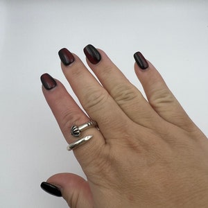 May include: A silver-colored ring in the shape of a nail, wrapped around a finger. The ring has a detailed head and a smooth, pointed end. The hand has dark red and black nail polish. The background is plain white.