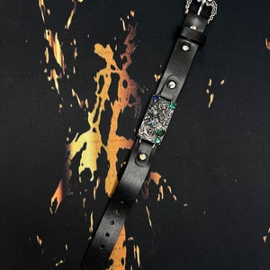 May include: A black leather bracelet with a silver buckle and a silver charm with green and blue gemstones. The bracelet is on a black background with gold splatter paint.