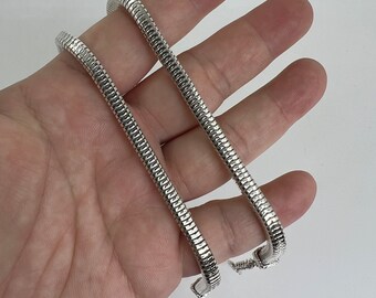 May include: A silver-colored, flexible bracelet with a snake-like design. The bracelet is made of small, interconnected metal segments, creating a textured surface. The bracelet is shown against a white background.