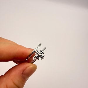 May include: A silver-colored ring with a floral design and a twisted band. The ring is held up against a white background. The ring has a small floral design in the center.