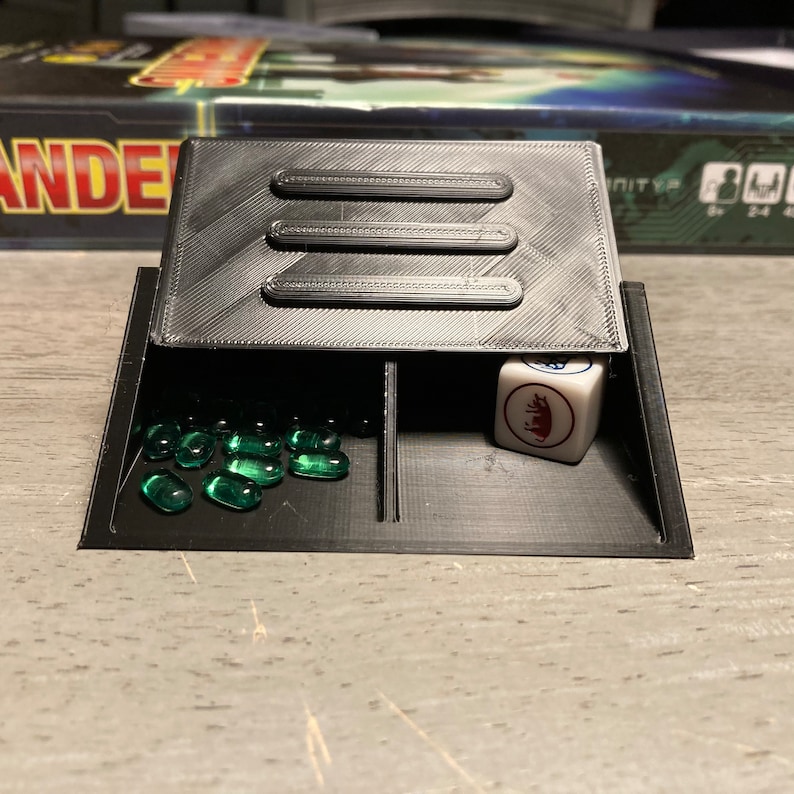 May include: A black plastic game tray with three compartments. The compartments contain green glass beads, a white die with a red bull symbol, and an empty compartment.