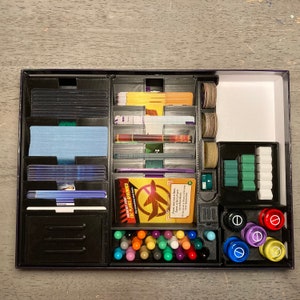 May include: A black and purple game organizer with compartments for cards, tokens, and dice. The organizer has a clear plastic lid and is filled with various game components, including a yellow and red card with the text "Direct First CDC Planes Grounded".