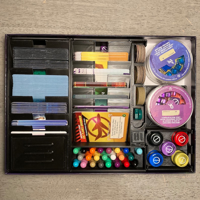 May include: A black board game organizer with multiple compartments for storing game pieces, cards, and tokens. The organizer has a variety of compartments, including a large compartment for cards, a smaller compartment for tokens, and a compartment for dice. The organizer is made of plastic and has a black finish.