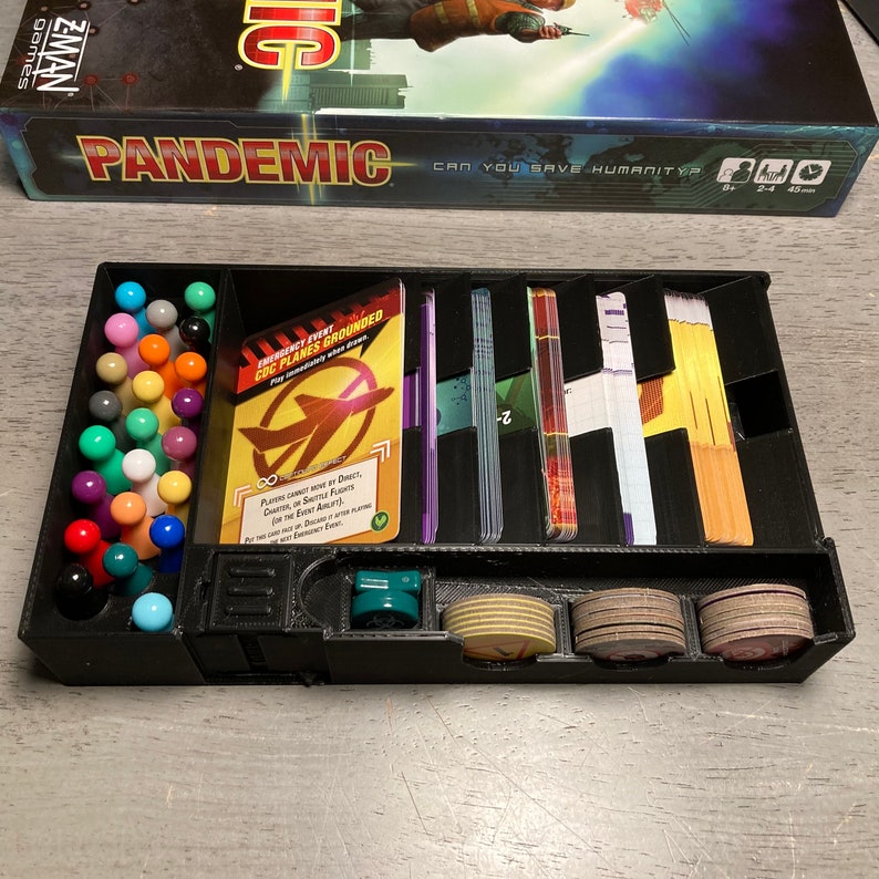 May include: A black plastic organizer for the Pandemic board game. The organizer has compartments for player pieces, cards, and tokens. The organizer also has a compartment for the game's rule book. The organizer is labeled with the game's title, "Pandemic".