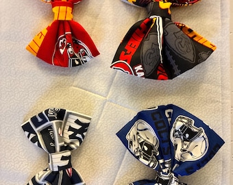 May include: Four fabric bow ties in red, black, blue, and navy. Each bow tie features a different sports team design, including the Kansas City Chiefs, New York Yankees, and Indianapolis Colts. The bow ties are arranged on a white quilted surface.