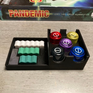 May include: A black plastic organizer for the board game Pandemic. The organizer has compartments for the game's colored cubes and player pieces. The game box is visible in the background with the text "Pandemic Can You Save Humanity?"